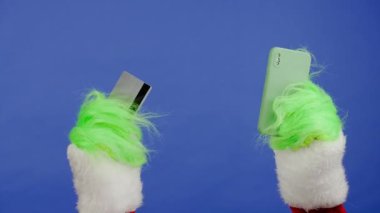 Grinchler yeşil saçlı eller, izole edilmiş mavi arka planda bir banka kartı ve akıllı telefon tutuyorlar. Hediye kaçıran kostüm. Noel ve Yeni Yıl kutlaması konsepti. Mavi ekran, krom anahtar