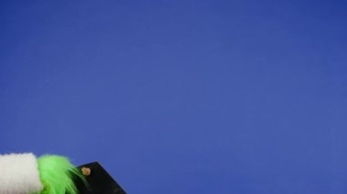 Grinchs 'in yeşil saçlı elinde mavi arka planda Mutlu Noeller yazan siyah bir tabela var. Hediye kaçıran kostüm. Yeni yıl kutlaması konsepti. Mavi ekran, krom anahtar.