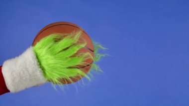Grinchs yeşil kıllı elleri mavi izole arka planda basketbol topunu tutuyor. Hediye hırsızı kostümü. Noel ve yeni yıl kutlaması konsepti. Mavi ekran, krom anahtar