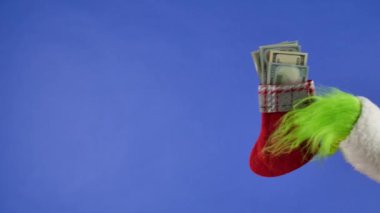 Grinchs 'in yeşil saçlı elleri mavi izole edilmiş arka planda dolar banknotları olan bir Noel çorabı tutuyor. Hediye Hırsızı Kostümü. Noel ve Yeni Yıl kutlaması konsepti. Mavi ekran, krom anahtar