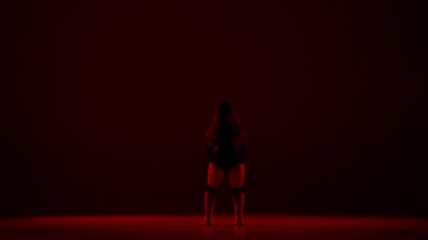 暗い背景のフレームには その上に赤い光が立っている若い女性が立っています ダンスムーブメントのスタイルをデモンストレーションします 彼女はセクシーでリズム的だ 彼女は開いた服を着ています — ストック動画