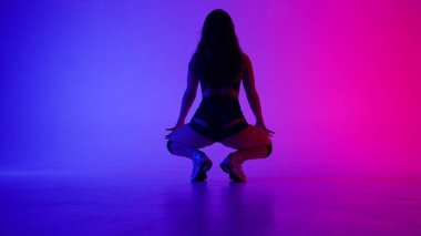 Çok renkli, koyu arkaplan, eğik çömelmiş bir kadın. Kıvırcık tarzında, açık kıyafetlerle bir dans gösterisi. Sırtı kameraya dönük duruyor, seksi, ritmik..