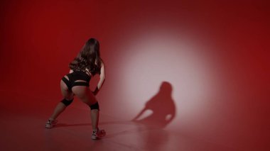 Çerçevede kırmızı arka planda genç bir kadın görünüyor. Spot ışıkları onun üzerinde parlıyor, gölgesi görünüyor. Kıvırcık tarzında bir dans hareketi gösteriyor. Açık giyinmiş, seksi, ritmik..