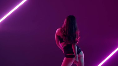 Mor renkli, neon arkaplanda parlak ışıklarla genç, narin bir kadın duruyor, twerk tarzında bir dans hareketi gösteriyor. Açık giyinmiş, seksi, ritmik, plastik..
