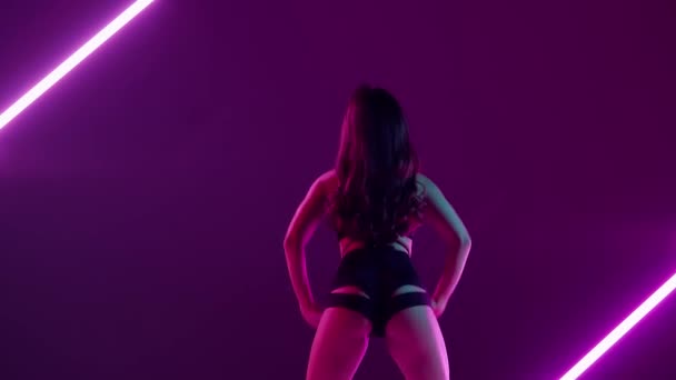 紫色のネオンバックグラウンドのフレームには 明るいライトが若くスレンダーな女性が立っている デモンは トゥーケのスタイルでダンスの動きを示しています 彼女はオープンな服 セクシー リズム プラスチックで服を着ています — ストック動画