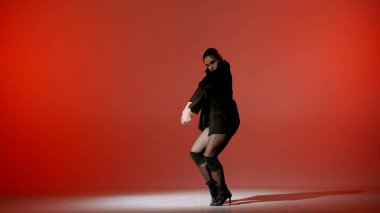 Kırmızı arka planda. Genç bayan, yüksek topuklara doğru dans hareketlerini gösteriyor. Üzerine bir spot ışığı parlıyor ve ona bir gölge veriyor, arka planın kenarları kararıyor. Seksi, ritmik..