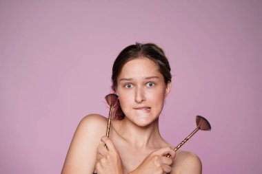 Dudağını ısıran ve makyaj fırçasıyla fırçalayan bir kadının portresi. Stüdyoda, açık tenli, çıplak makyajlı, pembe neon ışıkta pembe arka planda bir kadın. Yüz makyajı.