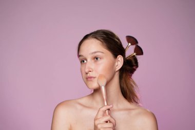 Makyaj fırçası kullanan bir kadın. Makyaj fırçasıyla tutturulmuş kadın saçı. Stüdyoda pembe ışıklı pembe arka planda bir kadın var. Yüz makyajı.