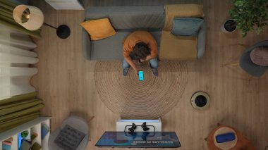 Akıllı ev teknolojisi yaratıcı konsepti. Dairedeki bir adamın portresi. Kanepede oturup elektrikli süpürgeye bakan, akıllı telefonu aletler için uygulamalı tutan video oyunu olan TV 'li adamın en iyi görüntüsü.