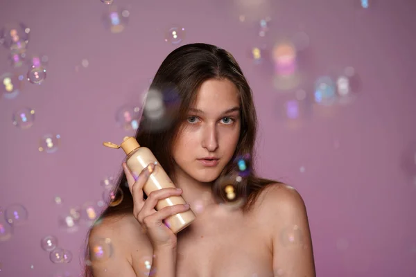 石鹸の泡に囲まれたピンクのバックグラウンドにスタジオで魅力的なセミナーの女性の肖像画 化粧品 クリーム ローション 血清のボトルを持っている女性 プロモーション — ストック写真