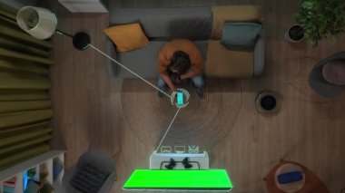 Akıllı ev ve modern teknoloji konsepti. Üst düzey bir adam kanepede oturuyor, akıllı telefondaki uygulamayı kullanarak ev aletlerini açıyor. Nesnelerin üzerindeki bilgi dijital görüntüler. Krom tuşlu yeşil ekranlı TV.