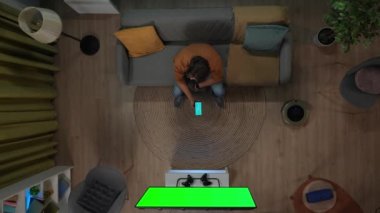 Akıllı ev ve modern teknoloji konsepti. Odada erkek portresi var. Kanepede oturan ve ev aletlerini çalıştıran adamın üst görüntüsü. Akıllı telefondaki uygulama, krom renkli yeşil ekranlı televizyon..