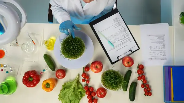 In the shot above, a scientist in a laboratory and a white coat is sitting. Around her on the table are vegetables and laboratory supplies. She is studying, examining, testing the greens. Top view.