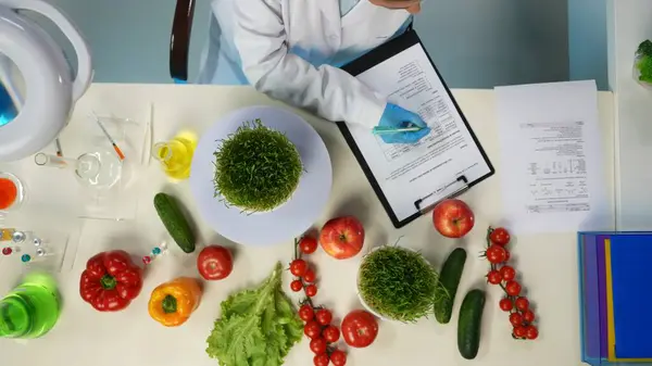 In the shot above, a scientist is sitting in a laboratory. Around her on the table are vegetables and laboratory supplies. She is studying, examining the greens. Shes writing something down. Top view.