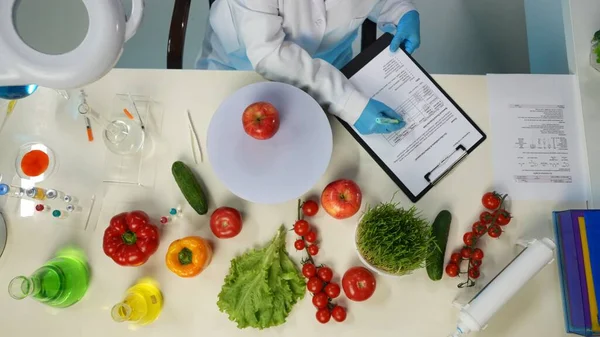 In the shot above, a scientist is sitting in a laboratory. Around her on the table are vegetables and laboratory supplies. She is studying, examining the apple. Shes writing something down. Top view.