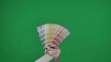 Yeşil arka planda çerçevelenmiş, kromakey. Manikürlü bir kadın eli tasarım için bir renk paleti tutar, renk seçimi için pastel renkler, gölgeler. Elini kaldırdı ve kameraya gösterdi..