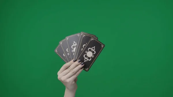 Framed on a green background, chromakey. A womans hand with a manicure holds playing tarot cards. She folded them like a fan and shows the outside with a beautiful pattern to the camera. Medium frame