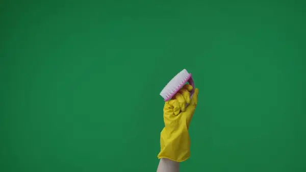 In the frame on a green background, a lame female hand in a yellow rubber glove holds and demonstrates a brush. Used for cleaning and cleaning items. Here can be your advertising. Medium frame.