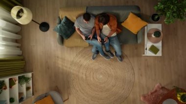 Evde homoseksüel bir çiftin en iyi görüntüsü. Kanepede oturuyorlar, ellerinde oyun kağıtlarıyla video oyunu oynuyorlar, eğleniyorlar ve birbirleriyle konuşuyorlar. LGBT, eşitlik, eğitim.
