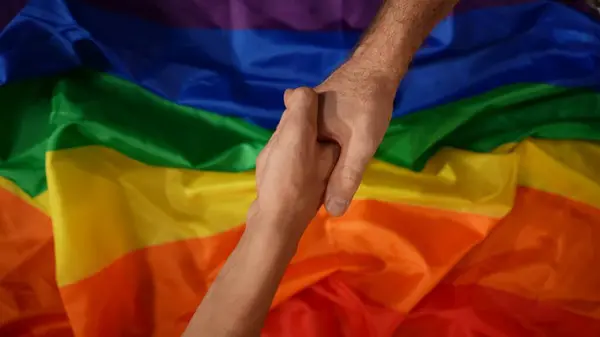 Üst görüntü, çerçevenin zıt köşelerinden el sıkışmaya çalışan iki erkeğin el sıkışmasını çekiyor. Arka planda LGBT bayrağı var. Eğitim içeriği, eşitlik, homoseksüellik, gurur.