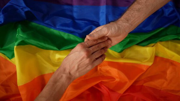 Top view detail shot capturing two male hands reaching for a each other from the opposite corners of the frame, touching gently. LGBT flag on the background. Educational content, equality, pride.