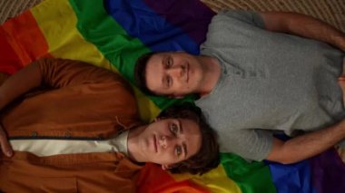 Homoseksüel, biseksüel, transseksüel bir çiftin LGBT bayrağına uzanmış kameraya gülümseyip, birbirlerine gülümseyerek aşk özgürlüğünü teşvik etmelerinin üst görüntüsü. Eğitim içeriği, gurur.