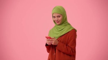 Orta ölçekli izole bir video, tesettür ve duvak takan çekici bir genç kadını çekiyor. Elinde akıllı bir telefon tutuyor, daktilo ediyor, kaydırıyor ve gülümsüyor. Reklamınıza, kültürünüze, çeşitliliğinize yer.