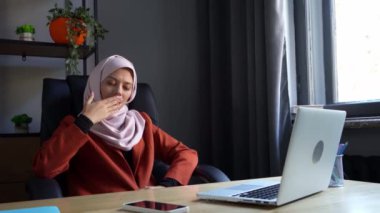 Laptopun önünde oturan tesettürlü çekici bir genç kadının orta boy videosu. Ağzını eliyle kapatarak esniyor. Kültürel çeşitlilik, iş ve eğitim, reklam.