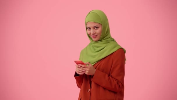 中規模の隔離されたビデオは ヒジャーブ ベールを身に着けている魅力的な若い女性をキャプチャします 彼女はスマートフォンを持ち タイピングし スクロールし 微笑んでいる 多様性のための場所 — ストック動画