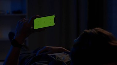 Geceleri kanepede uzanan ve elinde yeşil ekranlı bir akıllı telefon tutan bir adam. Adam video, film ya da dizi izliyor. Reklam alanı, işyeri