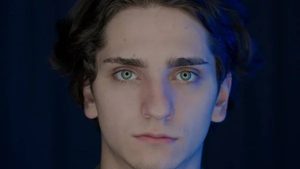 Portrait of a young man illuminated in blue color from a monitor screen on a black background close up. Man with tired eyes looking straight ahead. Concept of health and society
