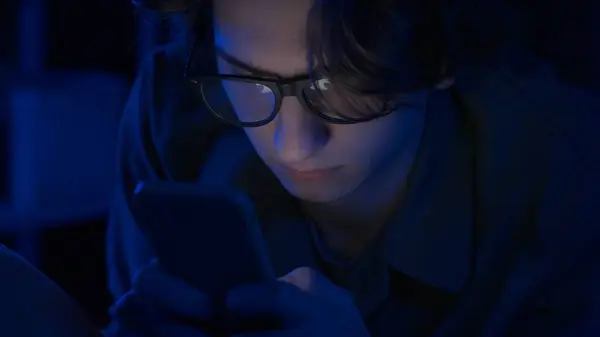 眼鏡をかけている若い男性は 夜に自宅でベッドに横たわるスマートフォンを使用しています 男性はテキストメッセージを送ったり オンラインゲームをしたり 写真やビデオを閲覧したりしています インターネット コミュニケーション — ストック写真