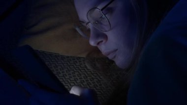 Gözlüklü genç bir kadın akıllı telefonuyla gece yatakta mesajlaşıyor. Teknoloji, internet, iletişim ve insan kavramı