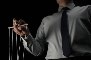 Çerçevede siyah bir arka planda elinde kukla tutan iplerle takım elbiseli bir adam duruyor. Birisini yöneten, yöneten bir iş adamını anlatıyor. Tiyatro performansı.