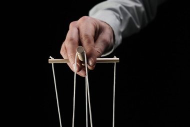Siyah bir arkaplana yakın çekimde. Kuklacı ipli bir aleti tutan bir erkek eli. Baskın bir adamı, bir işadamını, bir yöneticiyi gösteriyor. Tiyatro performansı, manipülasyon.