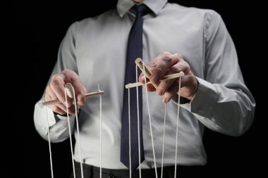 Çerçevede siyah bir arka planda elinde kukla tutan iplerle takım elbiseli bir adam duruyor. Birisini yöneten, yöneten bir iş adamını anlatıyor. Tiyatro performansı.