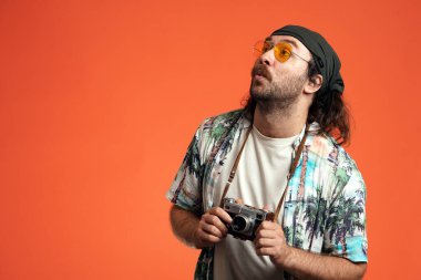 Retro kameralı erkek turist şaşkınlıkla bakıyor. Stüdyoda güneş gözlüklü bir erkek gezgin turuncu arka planda.