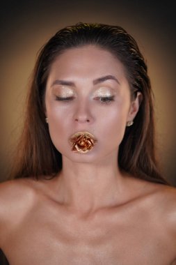 Güzellik ve moda yaratıcı reklam konsepti. Stüdyoda esmer kadın modelin portresi. Saç stili ve parlak göz makyajlı çekici bir kadın. Ağzında altın bir gül çiçeği tutuyor..