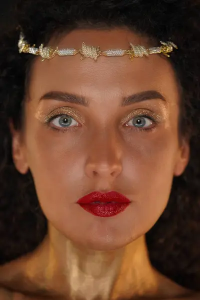 Beauty and glamour creative advertisement concept. Portrait of female model in studio. Close up face shot attractive girl with red lipstick wearing tiara, neck covered in golden paint looks at camera.