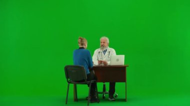 Sağlık ve tıbbi danışmanlık reklam konsepti. Krom anahtar yeşil ekranda yaşlı doktor portresi. Üniformalı kıdemli doktor masada oturmuş kadın hastalarla konuşuyor, tavsiyelerde bulunuyor..