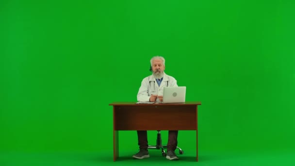 医療コンサルティング広告コンセプト クロマ グリーン スクリーンの老人の薬の肖像画 人とオンラインで話すヘッドセットラップトップのテーブルに座っている制服のシニアドクター — ストック動画