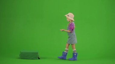 Bahçıvanlık ve tarım reklamları konsepti. Krom anahtar yeşil ekranda şapkalı ve lastik çizmeli bir kızın portresi. Küçük tatlı bahçıvan kız toprağı gübreledi..