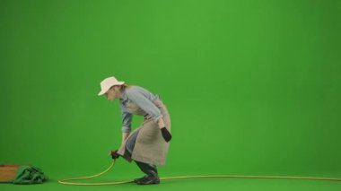 Bahçıvanlık ve tarım reklamları konsepti. Krom anahtar yeşil ekranda önlüklü kadın portresi ve lastik çizmeler. Kadın bahçıvan aletleri kontrol ediyor ve çiçeklerini bahçe hortumuyla suluyor..