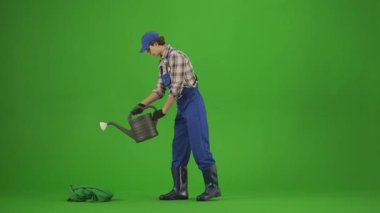 Bahçıvanlık ve tarım reklamları konsepti. Tulumlu erkek portresi ve krom anahtar yeşil ekranda lastik çizmeler. Erkek bahçıvan, bitki yataklarını bir su bidonuyla suluyor..