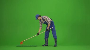 Bahçıvanlık ve tarım reklamları konsepti. Tulumlu erkek portresi ve krom anahtar yeşil ekranda lastik çizmeler. Bahçıvan adam gevşer ve zemini tırmıkla düzleştirir, sonuçlara bakar..