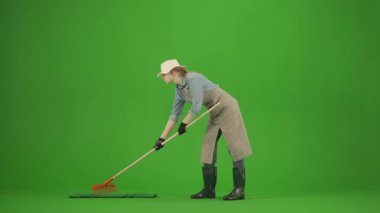 Bahçıvanlık ve tarım reklamları konsepti. Krom anahtar yeşil ekranda önlüklü kadın portresi ve lastik çizmeler. Kadın bahçıvan zemini tırmıkla düzleştiriyor, olumlu bir ifadeyle bakıyor.