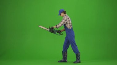 Bahçıvanlık ve tarım reklamları konsepti. Tulumlu erkek portresi ve krom anahtar yeşil ekranda lastik çizmeler. Erkek bahçıvan modern testere ile odun kesiyor, pozitif ifade..