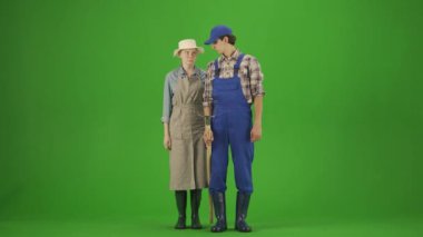 Bahçıvanlık ve tarım reklamları konsepti. Krom anahtar yeşil ekranda sıradan ve lastik çizmeli bir çift portresi. Kadın ve erkek bahçıvanlar yabayla poz veriyor, el ele tutuşup kameraya bakıyor.