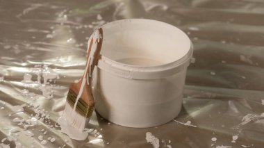 Çerçeve, bir kova boyanın üzerinde dikey duran beyaz boyalı bir fırçayı gösteriyor. Yağ beziyle kaplı bir zeminde duruyorlar. Boya damlıyor, su birikintisi oluşturuyor..