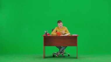 Modern çocuk okulu ve eğlence zamanı reklam konsepti. Krom anahtar yeşil ekrandaki çocuk portresi. Okul çocuğu masada oturuyor, okul ödevlerini yapıyor, deftere yazıyor, esnemekten yorulmuş..